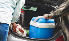 Geöffneter Kofferraum eines Autos, zwei Personen laden Taschen und eine blaue Kühlbox ein