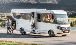 Niesmann+Bischoff iSmove (2024) parkt auf einer asphaltierten Fläche, eine Person lädt ein Fahrrad in die Heckgarage, eine andere Person steht in der Eingangstür.