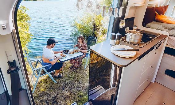 Das Bild zeigt die Küche des Bürstner Lineo C 590 (2024). Die Schiebetüre ist geöffnet, vor dem Fahrzeug sitzen zwei Personen an einem Campingtisch und essen.