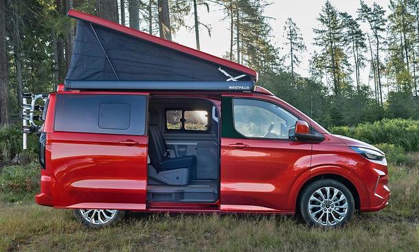 Ford Transit Custom Nugget (2024) auf einer Wiese, dahinter Wald, mit aufgestelltem Dach und offener Schiebetür.