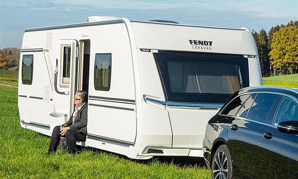 Fendt Caravan Apero 560 SKM (2024) hängt an einem Auto und parkt auf einer Wiese. In der Eingangstür zum Caravan sitzt eine Person.