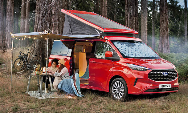 Ford Transit Custom Nugget (2024) auf einer Wiese, dahinter Wald, mit aufgebauter Markise und aufgestelltem Dach. Die Schiebetür ist geöffnet, zwei Personen sitzen unter der Markise und Campingstühlen.