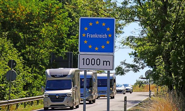 Das Bild zeigt ein Schild mit der Aufschrift "Frankreich, 1000 m" nahe der Grenze zu Frankreich. Auf der Straße fahren drei Wohnmobile in Richtung Kamera.