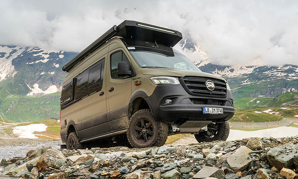 Sam the Van (2025) stehend, von schräg vorne rechts. Das Fahrzeug steht mit den Hinterrädern in einem Bach, die Vorderräder sind am Ufer. Im Hintergrund sind wolkenbehangene Berge zu sehen.