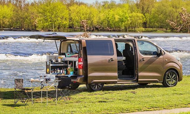 Toyota-Campingzubehör; Toyota Proace Verso, stehend, mit offener Heckklappe und Schiebetüre. Im Van befindet sich eine Campingbox, davor steht ein Tisch mit Klappstühlen.